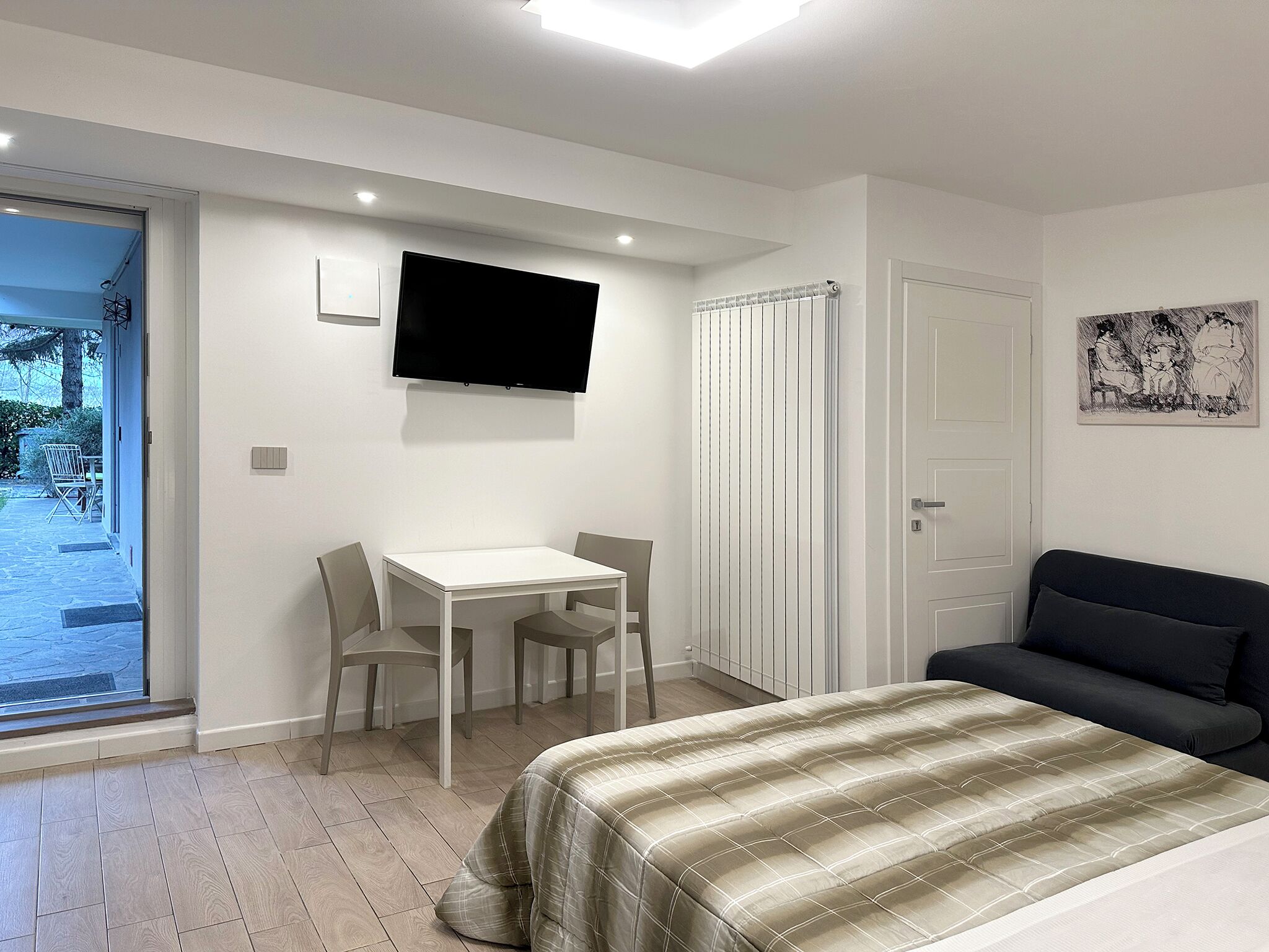 A bedroom with a bed, a TV mounted on the wall, a small table with chairs, and a door leading to a balcony.