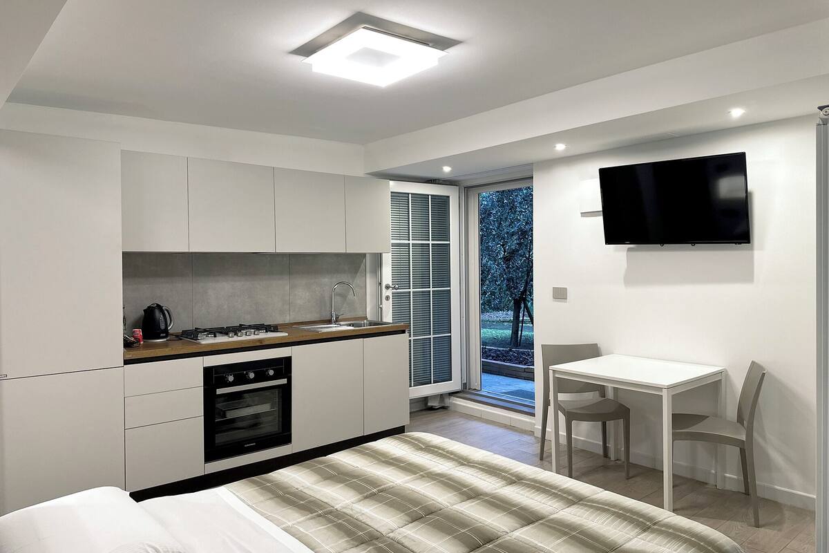 Quarto moderno com cama, pequeno espaço de cozinha, TV e janela com persianas.