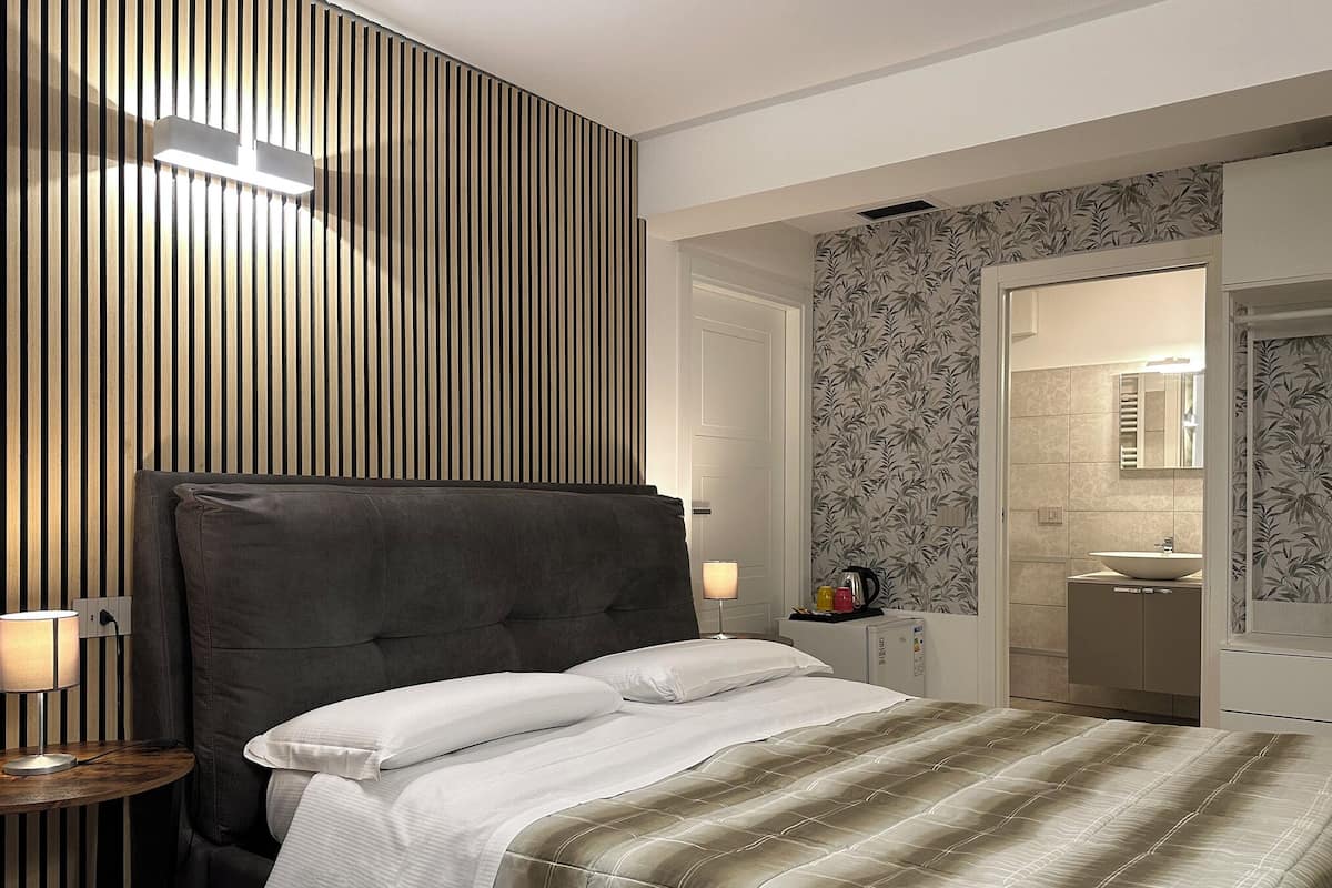 Quarto de hotel moderno com uma cama grande, uma mesinha de cabeceira com abajur, parede com ripas de madeira, banheiro com pia e espelho, e papel de parede estampado.