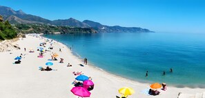 Una playa con gente tomando el sol y nadando, cielo despejado y montañas al fondo.