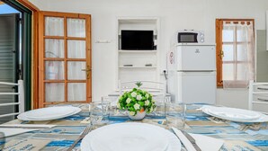 Une table à manger dressée avec des assiettes, des couverts et un arrangement floral au centre, avec un téléviseur et un réfrigérateur en arrière-plan.