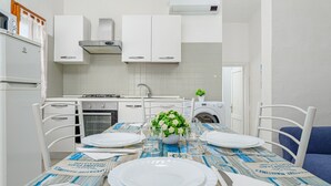 Une cuisine moderne avec des armoires blanches, un four encastré et un coin repas agrémenté d’une table dressée avec de l’assiette blanche et une nappe bleue.