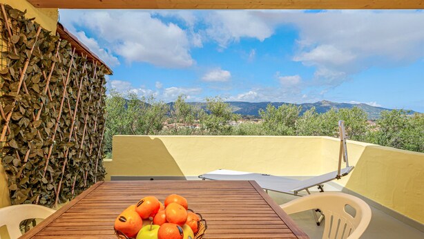 Une table en bois sur laquelle se trouve un panier de fruits, une chaise blanche et une vue sur un paysage montagneux.