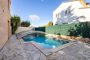 Ein Pool mit gewölbter Kante, Steinfliesen, einem weißen Gebäude und einer grünen Hecke.