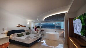 A modern bedroom with a large bed, a bathtub, and a city view. - Relais Manfredi (Castellammare di Stabia)