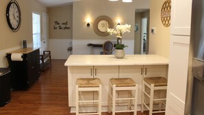 Una cocina con una isla central, taburetes de bar, un reloj y una pared con espejo y luces decorativas.