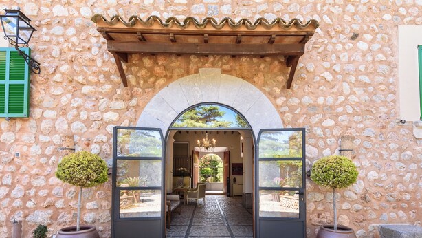 Une entrée en pierre, avec une porte en arc de cercle, deux arbres taillés en forme de cône placés dans des pots, et un sol carrelé menant à un espace intérieur.