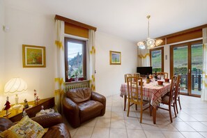 Een woonkamer met een bruine leren bank, een eettafel met stoelen, een radiator en een raam met gordijnen.