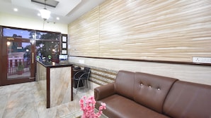 A reception area with a brown leather sofa, a glass coffee table with a pink flower arrangement, a reception desk, and a view through a glass door. - Hotel New Palace (Dalhousie)