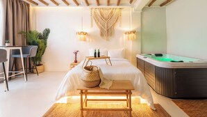 A modern hotel room with a bed, a hot tub, a desk with chairs, and a large plant.