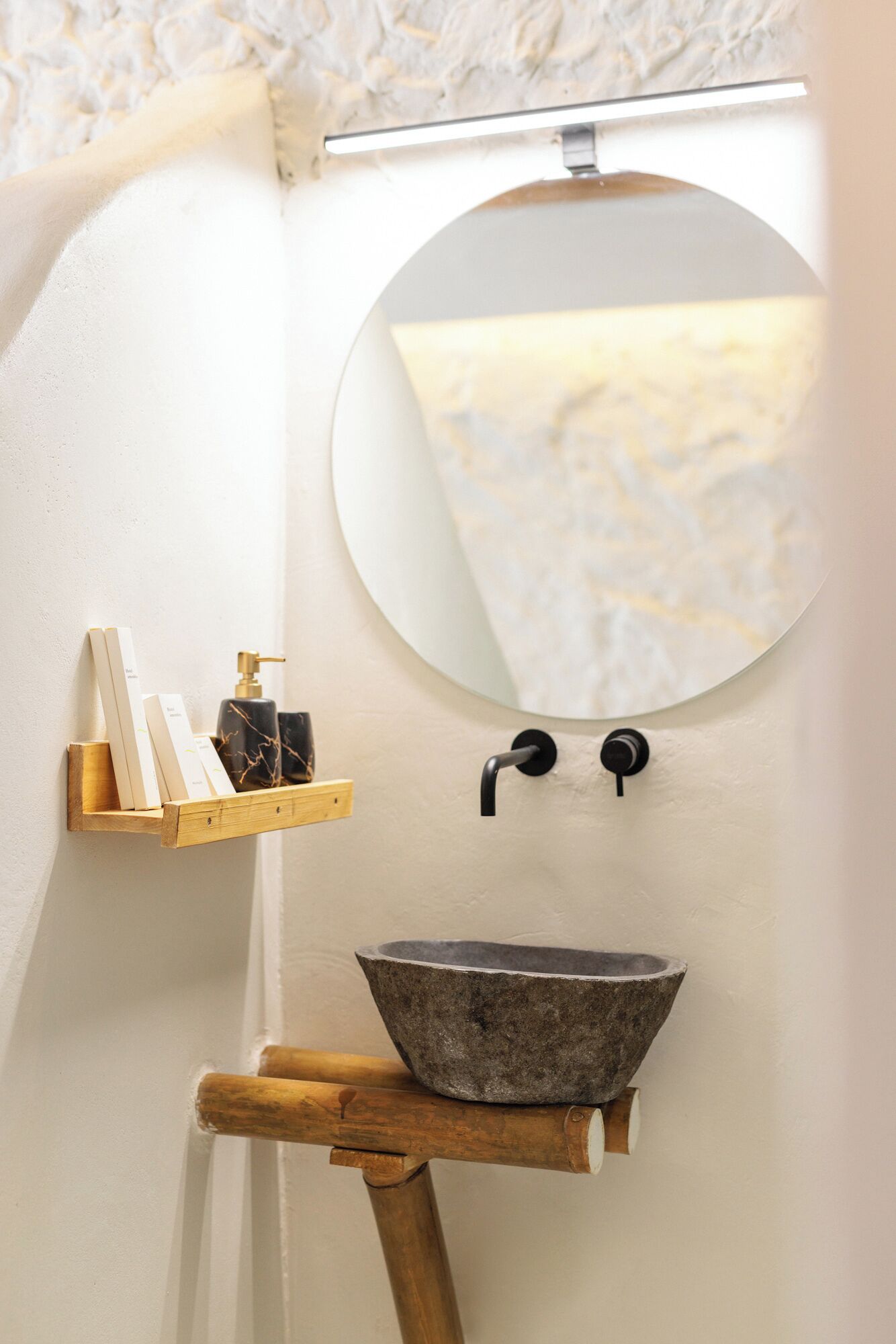 A bathroom with a round mirror, a stone sink, and a wooden shelf with toiletries.