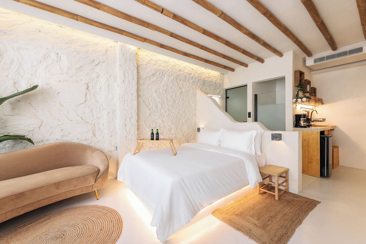 A modern bedroom with a bed, a sofa, a small table with bottles, and a kitchenette area.