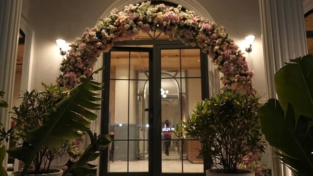 Entrada de hotel con un arco floral, plantas en macetas y un mostrador de recepción visible a través de las puertas de cristal.