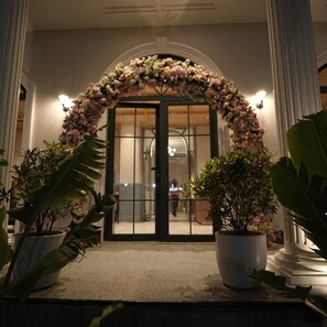Entrada de hotel con un arco floral, plantas en macetas y un mostrador de recepción visible a través de las puertas de cristal.