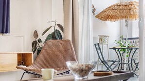 Una habitación con una silla de mimbre, una mesa de madera con un tazón y una taza, una planta y vistas a un espacio exterior con una mesa y sillas.