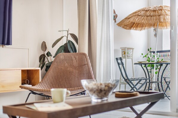 Une pièce avec un fauteuil en osier, une table en bois sur laquelle se trouvent un bol et une tasse, une plante, et une vue sur un espace extérieur aménagé avec une table et des chaises.
