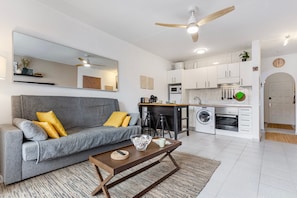 Un espace de vie compact comprenant un canapé gris, une table basse, un coin cuisine avec un four à micro-ondes et un lave-linge.