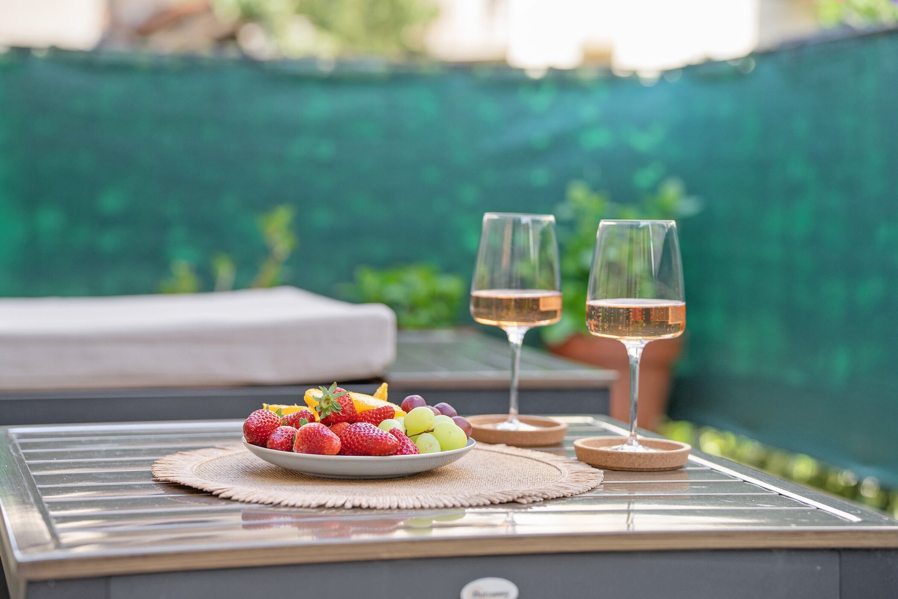 A table with a fruit bowl and two glasses of wine, set against a green background.