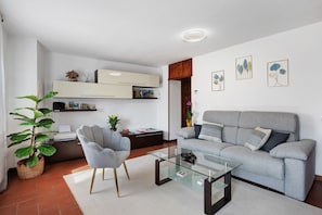 Una sala de estar moderna con un sofá gris, una mesa de centro de vidrio, un sillón gris, una estantería y una planta en maceta.