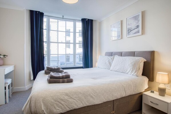 A hotel room with a bed, bedside table, lamp, and a window with curtains. - Skokholm Apartment - 3 Bedroom Apartment - Tenby (Tenby)