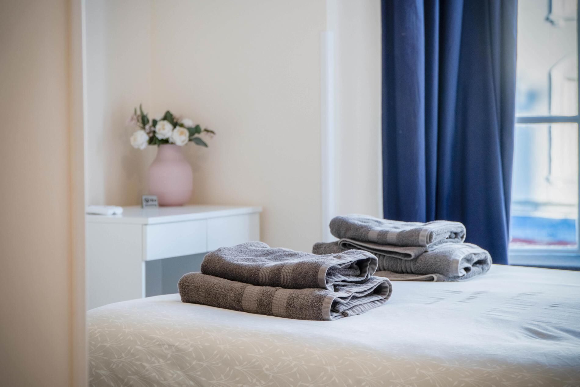 Una cama bien tendida con toallas dobladas, una mesita de noche con un florero y una ventana con cortinas.