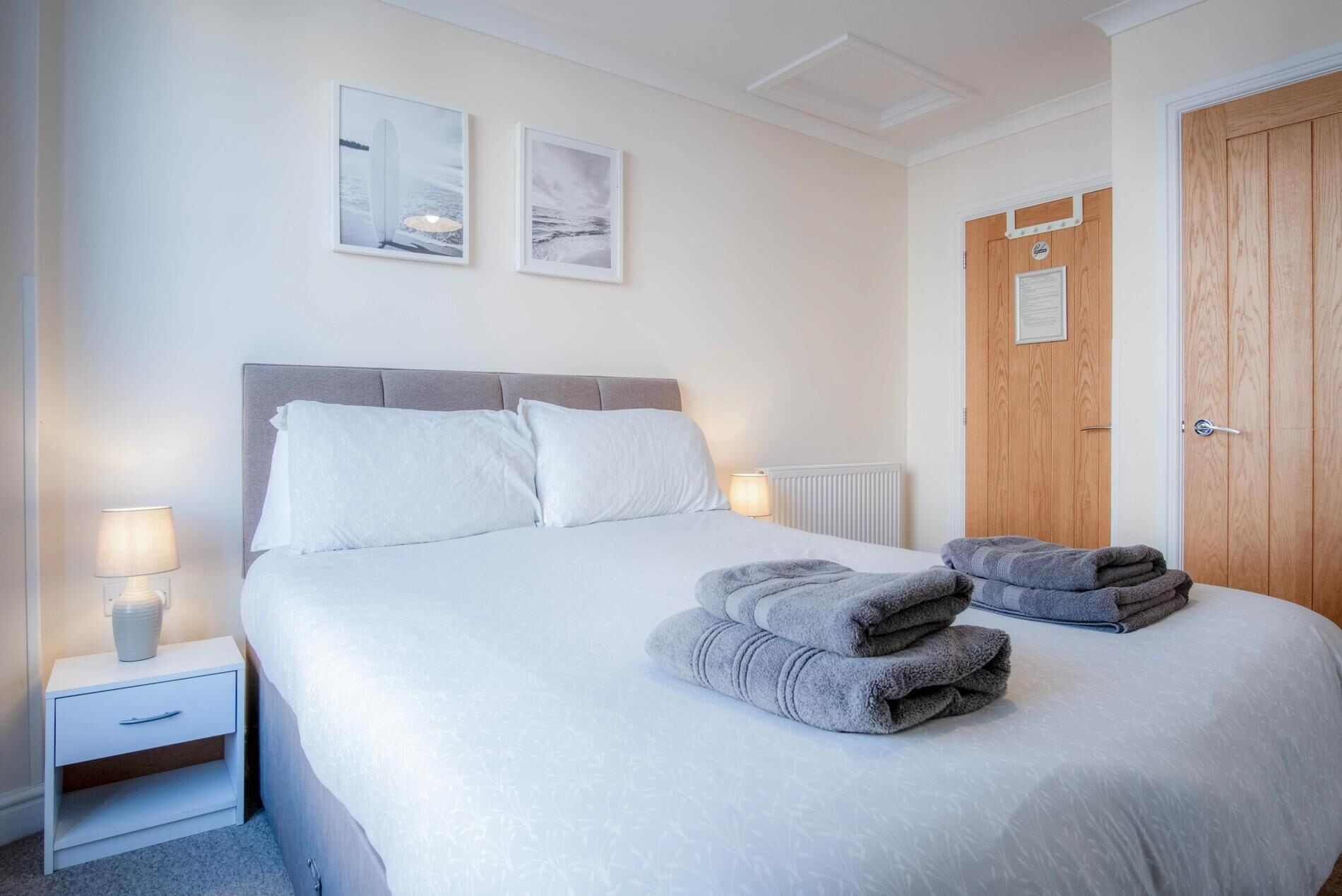 Un lit bien fait, avec du linge de lit blanc et deux serviettes pliées posées sur le lit. On aperçoit une table de chevet avec une lampe et un petit objet. La pièce est dotée d’une porte en bois et d’un tableau encadré accroché au mur.