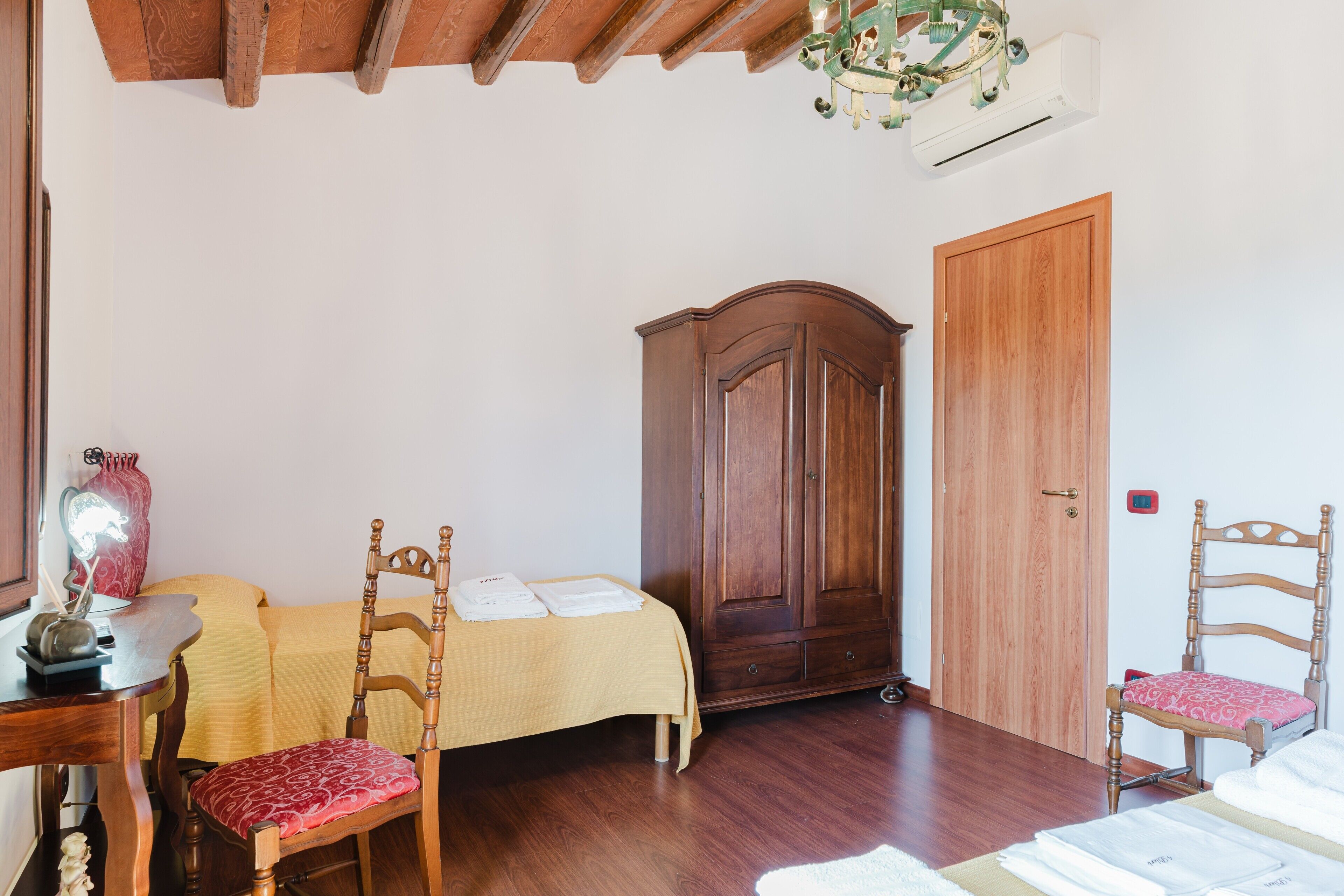 Una habitación con una cama, dos sillas de madera, un armario de madera, una mesa con un florero y un ventilador de techo.