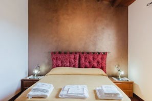 Un lit bien fait, recouvert d’une couvre-lit beige, avec des rideaux à motifs rouges et deux tables de chevet sur lesquelles sont posées des lampes et des serviettes pliées.