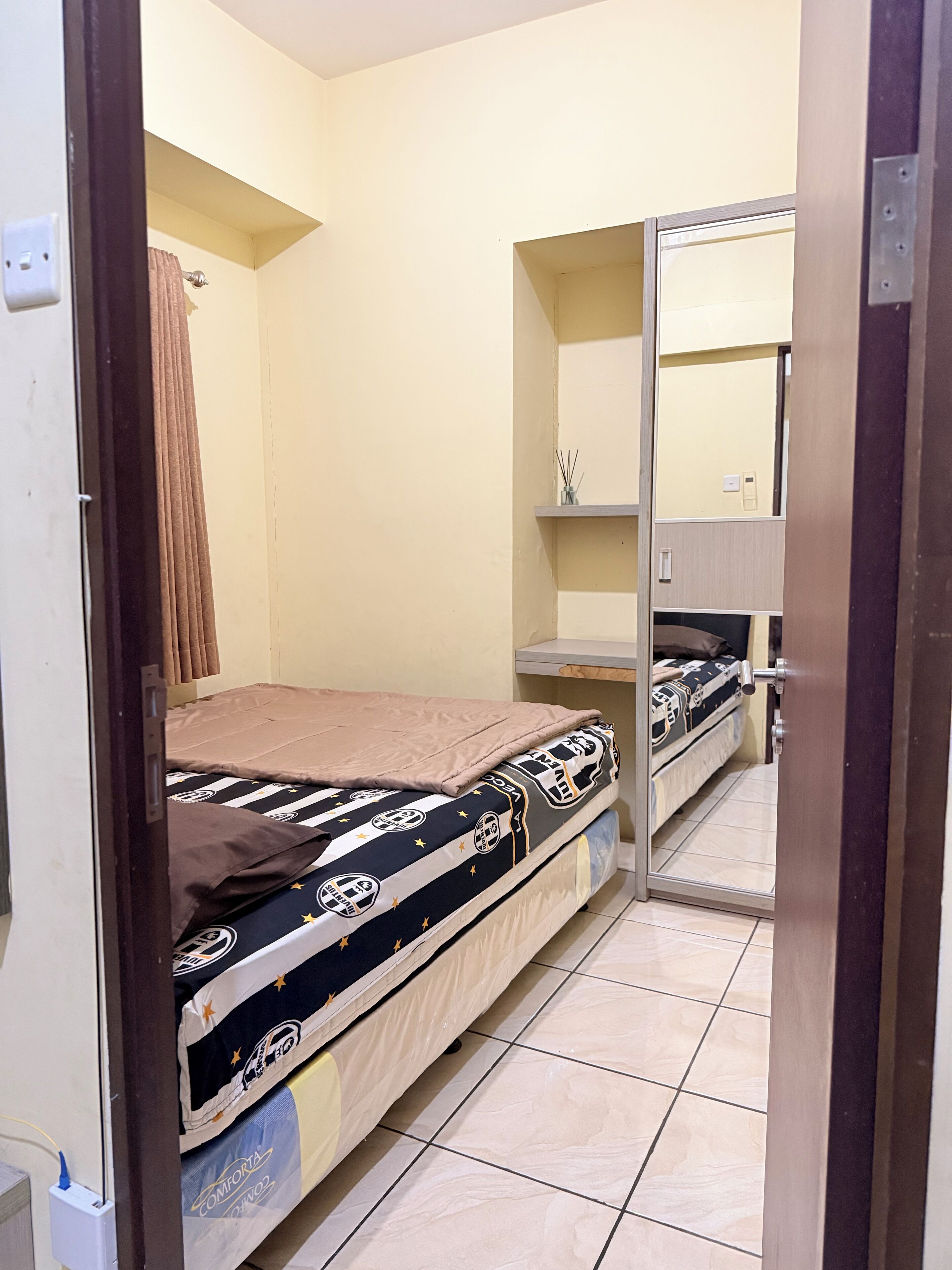 A small, compact room with a single bed, a mirror, and a shelf with toiletries.