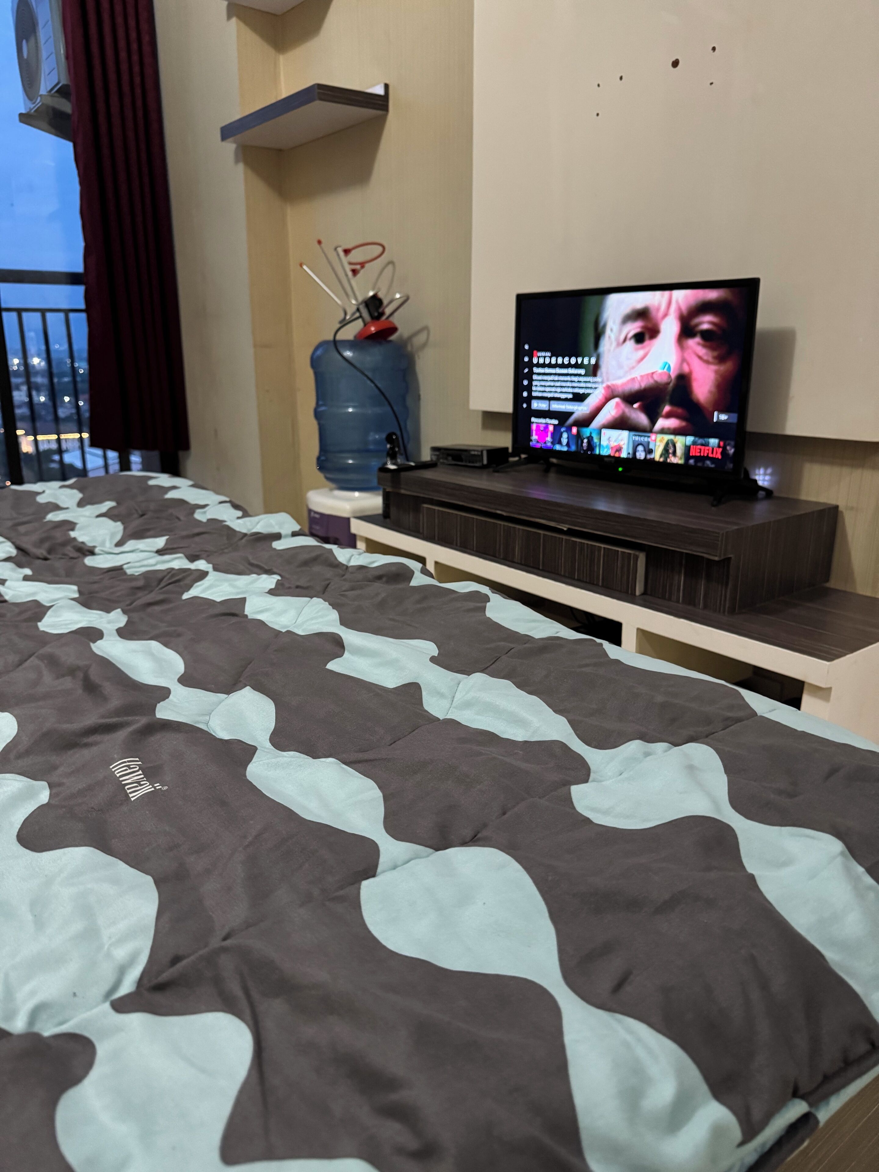 A bedroom with a bed, a TV on a wooden stand, a water dispenser, and a view of the outside.