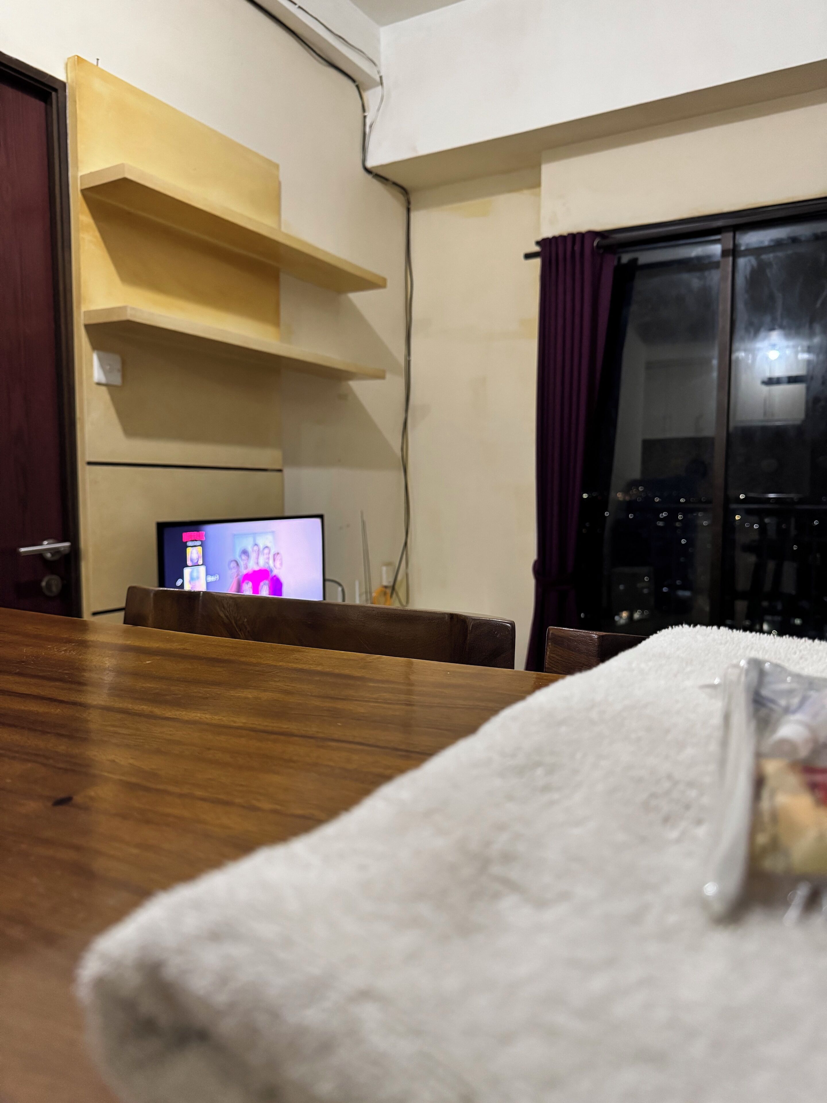 Ein Zimmer mit einem Holzesstisch, einem Fernseher auf einem Schrank und einem Fenster mit violetten Vorhängen.