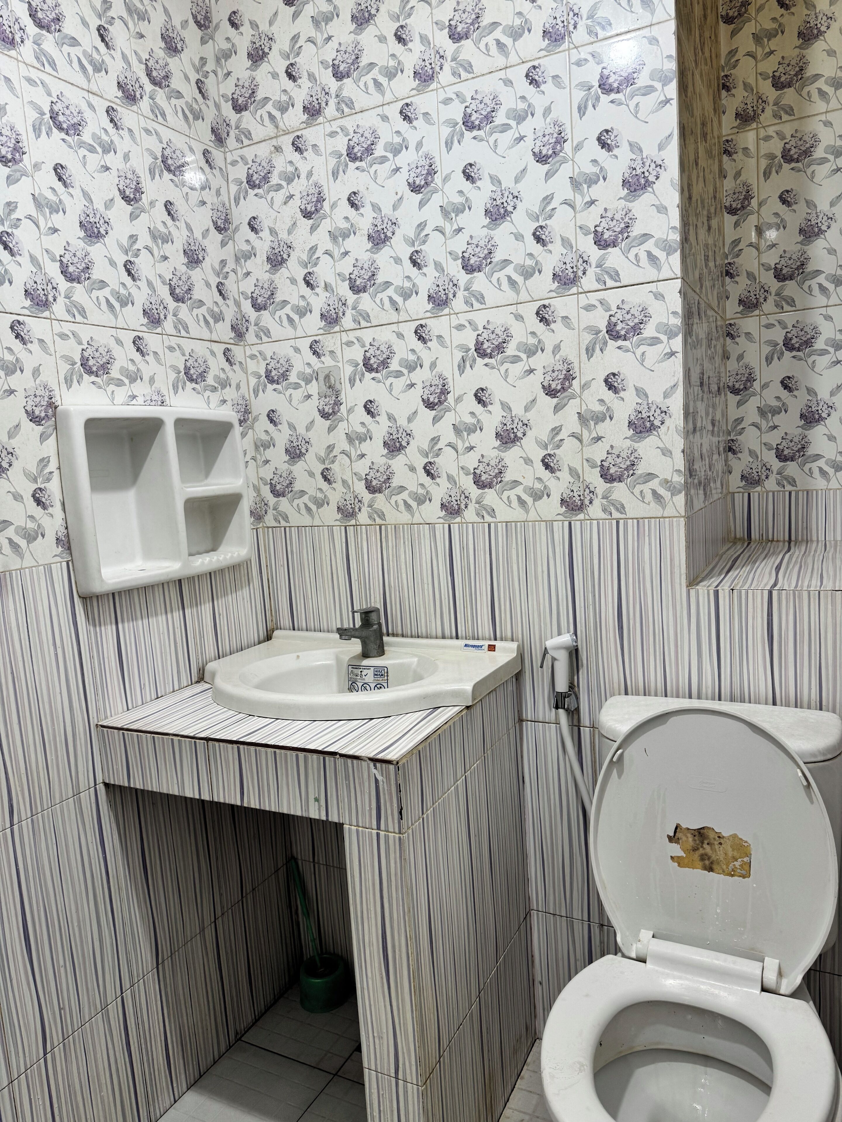 Une salle de bain avec des murs recouverts de carreaux à rayures, un lavabo blanc avec un distributeur de savon et des toilettes dont l’abattant est endommagé.