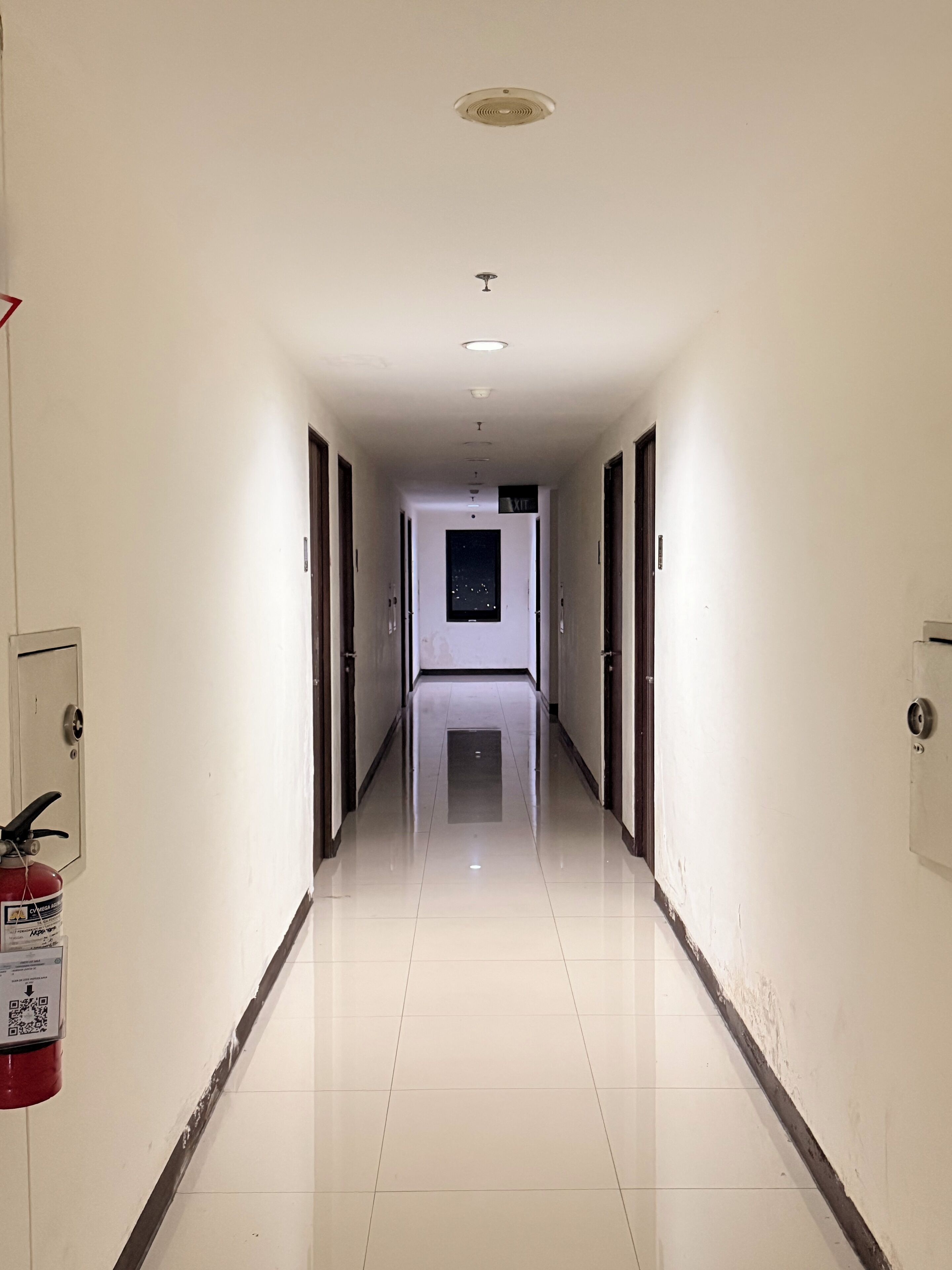 Un long couloir étroit aux murs blancs et au sol carrelé, qui mène à une porte au bout.