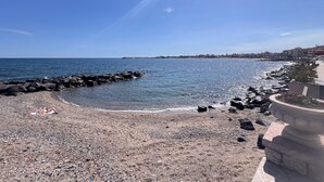 En strand med småsten och en vågbrytare av sten, klarblå himmel och byggnader i fjärran.