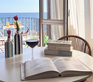 A table with an open book, a wine glass, and two vases with flowers. There are books and a fruit bowl on the table. - Tysandros by the Sea (Giardini Naxos)