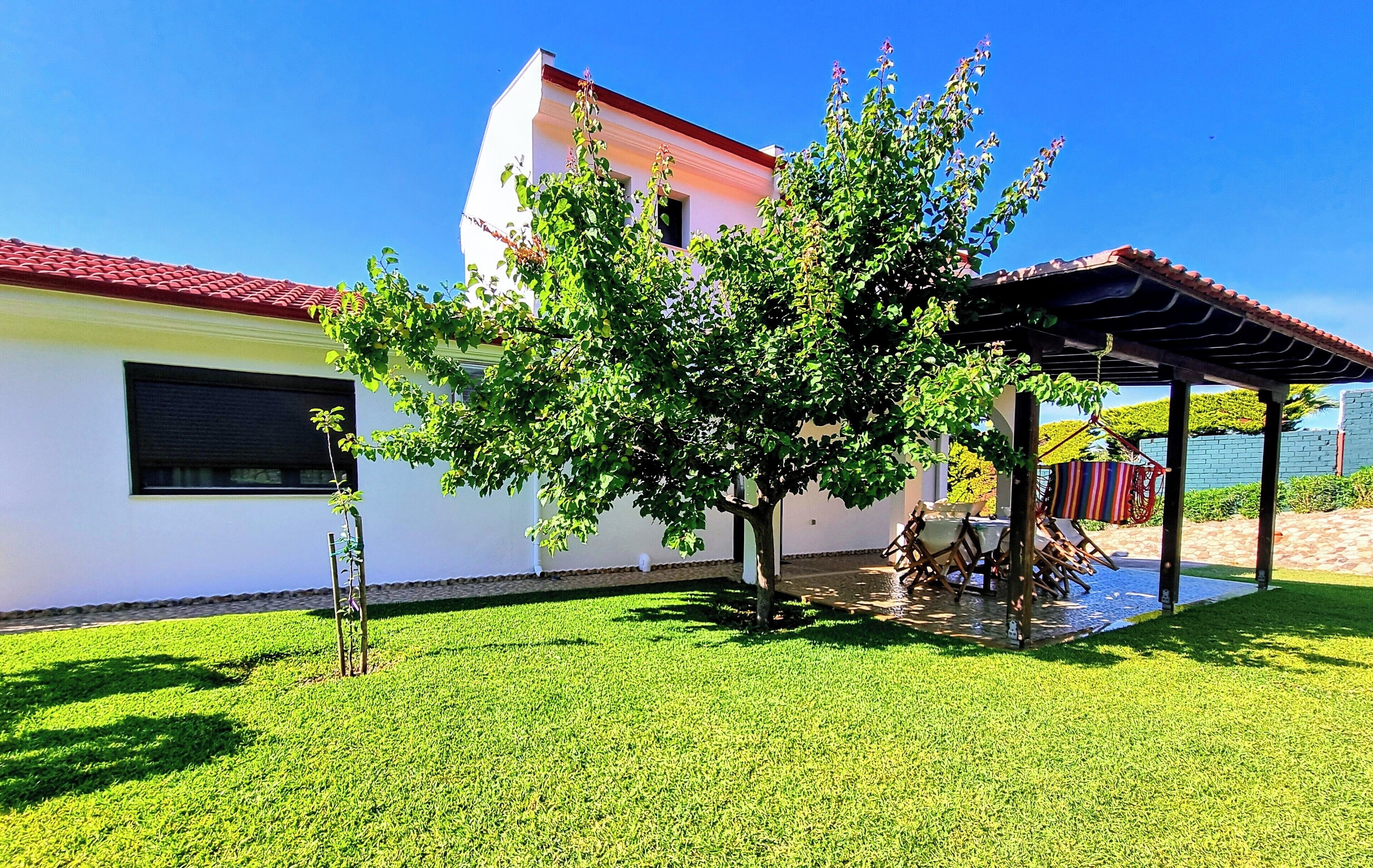 A white building with a red roof, a tree in the front yard, and a covered patio area with outdoor furniture.