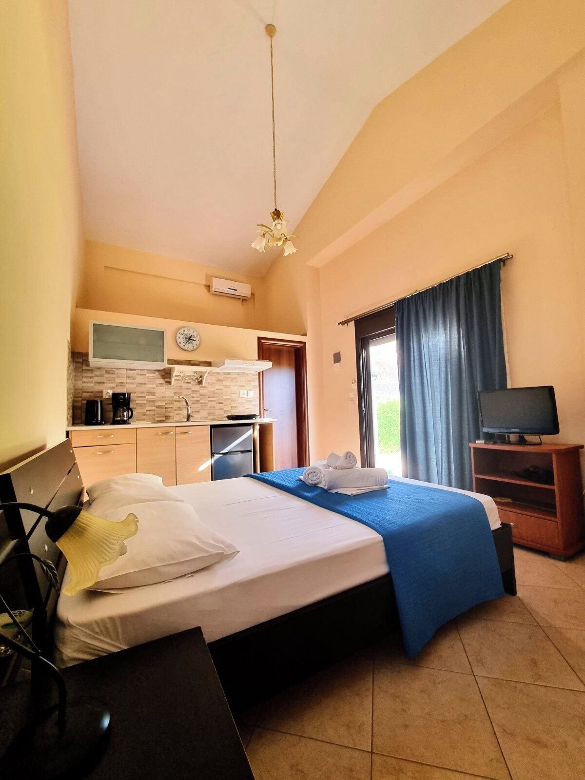 A hotel room with a bed, a desk, a television, a kitchenette, and a bathroom visible through an open door.