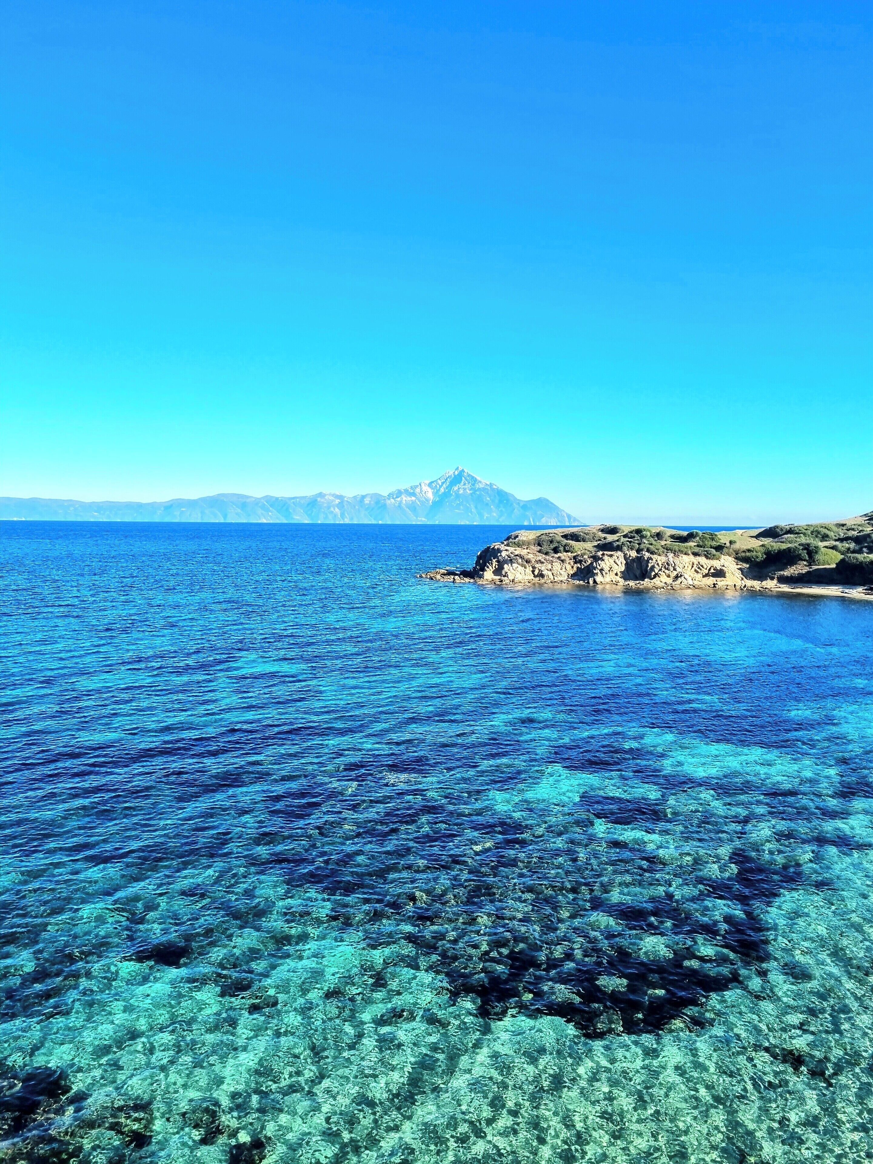 A coastal landscape with clear blue waters, rocky terrain, and distant mountains.