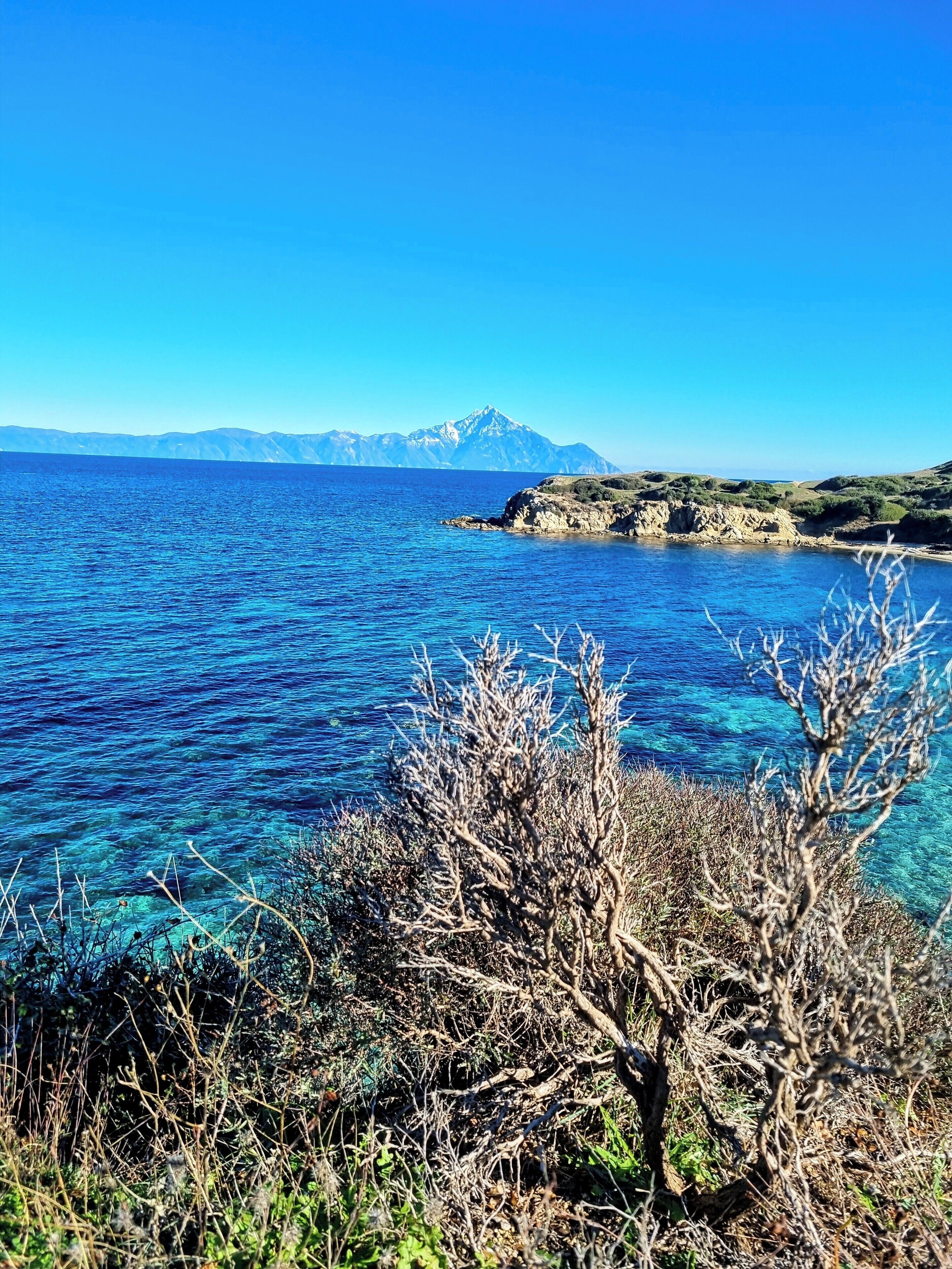 A coastal landscape with clear blue sea, rocky terrain, and a mountain in the distance.