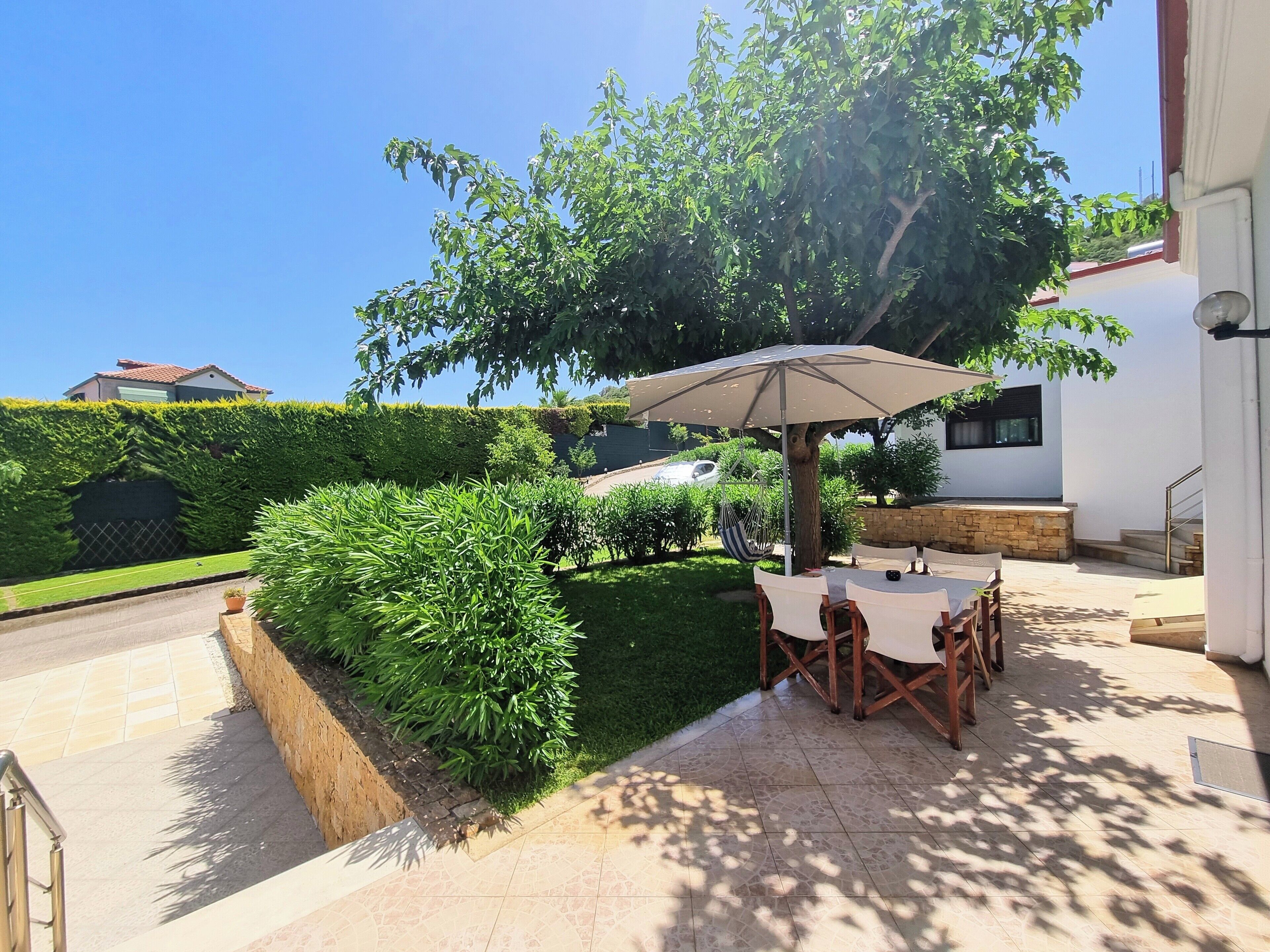 An outdoor patio area with a table and chairs under a large umbrella, surrounded by well-maintained hedges and a lawn.