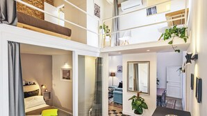 Un appartement moderne avec un sol à carreaux, un lit avec des oreillers, un coin repas avec un saladier et un escalier menant à une mezzanine.