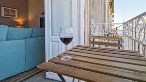 Une table en bois avec un verre de vin rouge, un canapé de couleur turquoise et une vue sur le paysage urbain, visible à travers la rambarde d’un balcon.