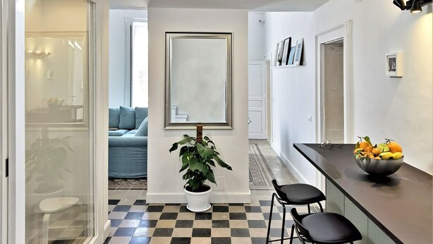 Un espace de vie moderne et ouvert, avec un sol à carreaux, un coin repas doté de tabourets de bar, une plante en pot placée au centre et un escalier menant à un niveau supérieur.