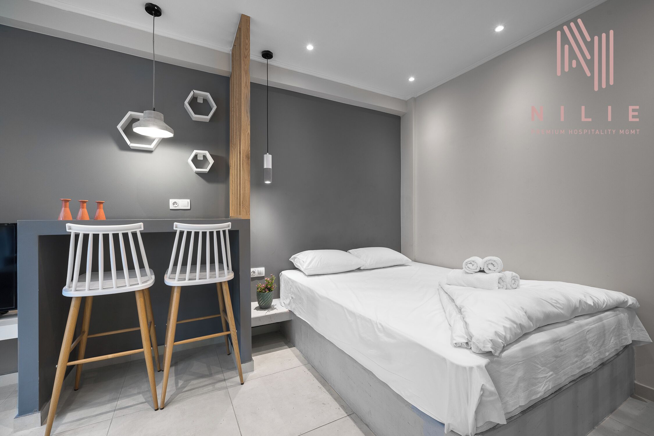 A modern hotel room with a bed, two white bar stools, a small table, and a wall-mounted light fixture.