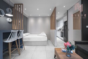 Una habitación de hotel moderna con una cama, dos taburetes blancos, un separador de madera, una mesa de comedor con un florero y un sofá.