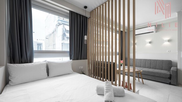 A modern hotel room with a large bed, grey sofa, and a view of the city. - Bonsai Studios & Apartments, Nilie Hospitality MGMT (Thessaloniki)
