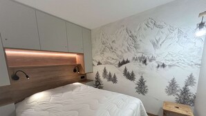 A bedroom with a bed, a headboard with integrated lighting, a wall mural of a mountainous landscape, and a small wooden stool.