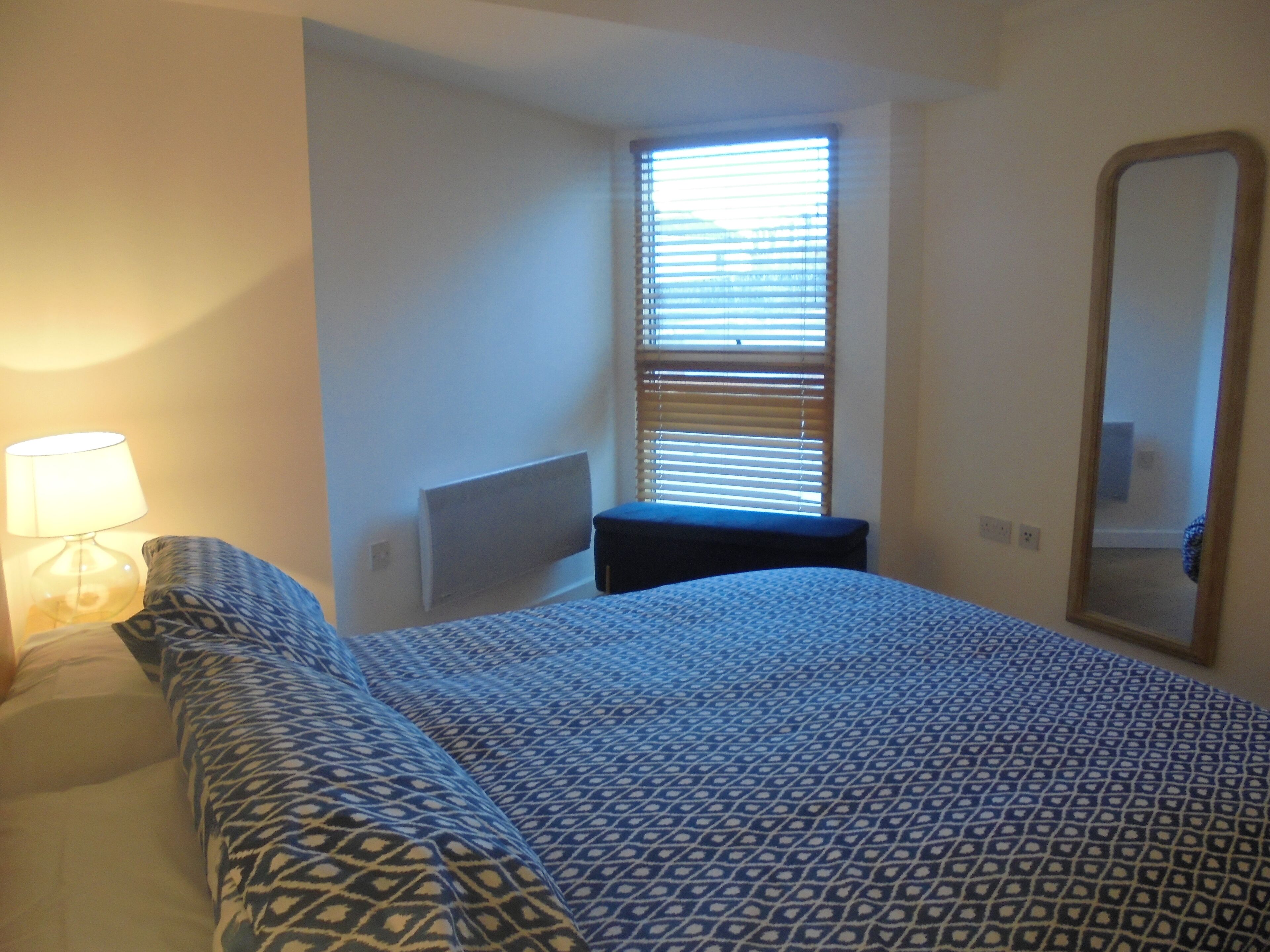 A bedroom with a bed, a bedside lamp, a window with blinds, a small bench, and a mirror.