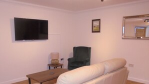 A living room with a white sofa, a television mounted on the wall, a chair, and a small table with a lamp.