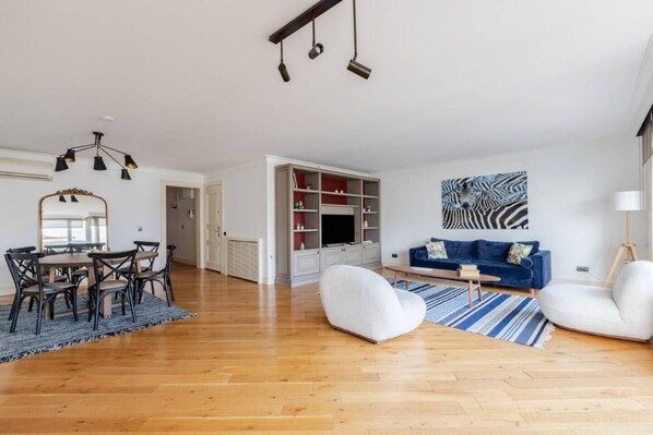 Een ruime woonkamer met houten vloer, een blauwe bank, een witte fauteuil, een eettafel met stoelen en een televisie in een inbouwmeubel.
